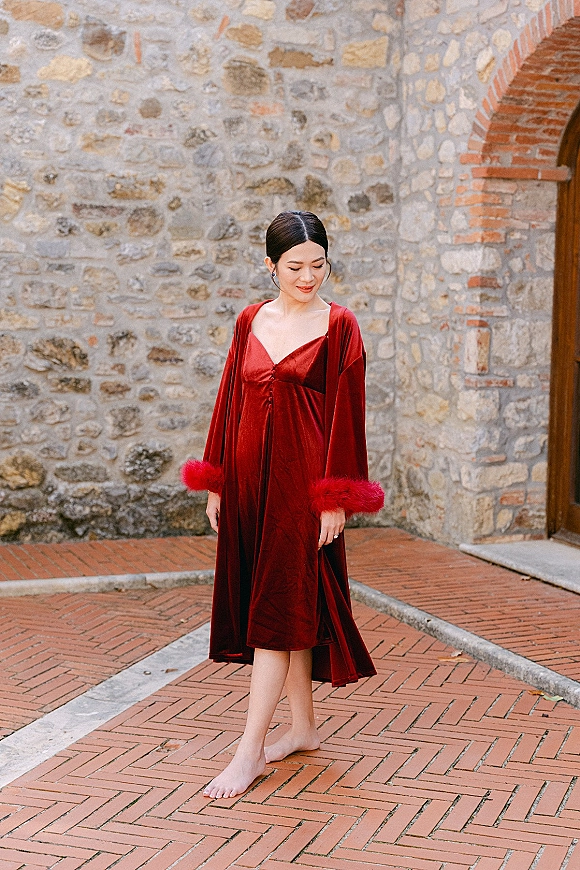 Bridesmaid portrait in a burgundy bridesmaid dress and matching robe with feather cuffs, standing barefoot by a stone wall and brick archway
