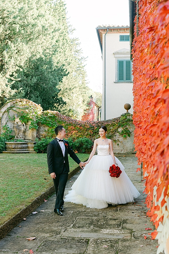 Couple portrait of bride and groom holding hands, bride in strapless ball gown with red rose bouquet walking past ivy villa wall and fountain
