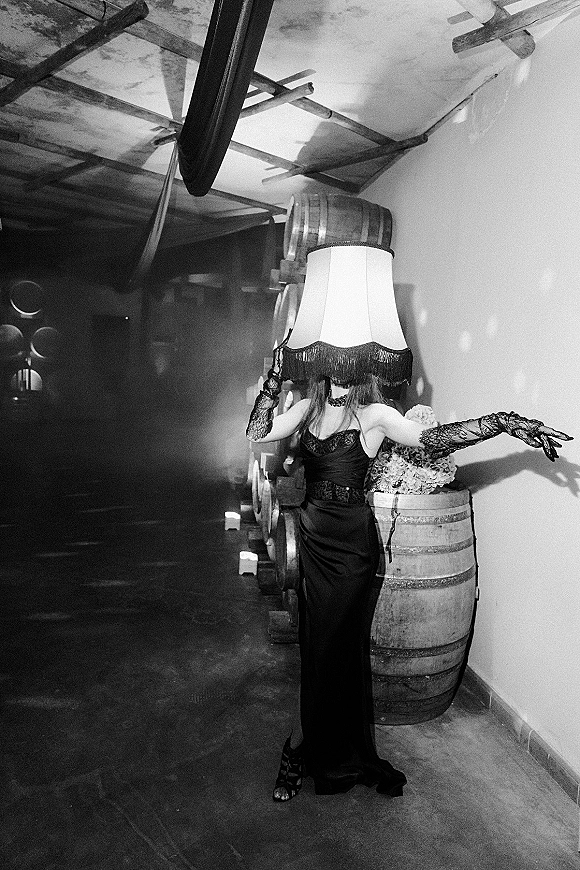 Bridal portrait in a black wedding dress with lace gloves and a lampshade headpiece, posing by candlelit wine barrels in a cellar hallway