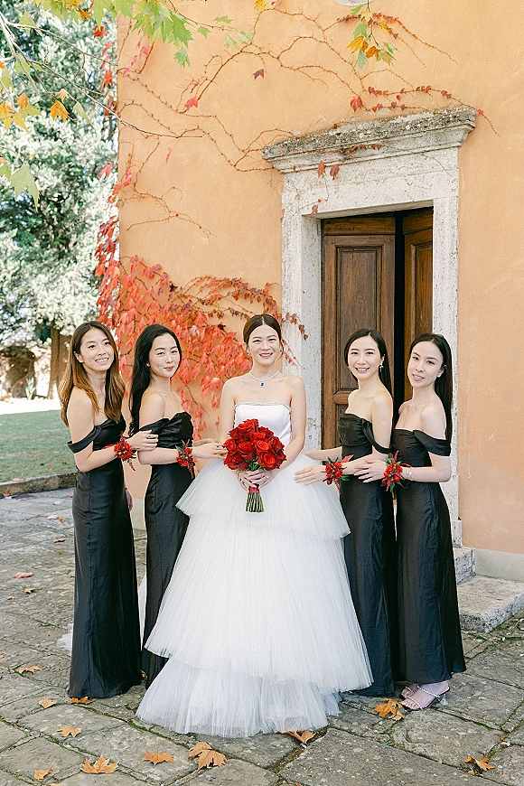 Bride with bridesmaids portrait showing her in a white strapless ball gown holding red roses beside black satin dresses by an ivy stucco wall doorway