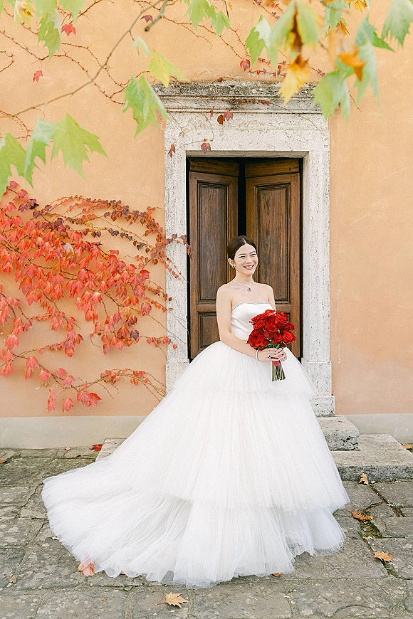Bridal portrait of a bride in a strapless ball gown holding a red rose bouquet in a stone doorway with ivy on a stucco wall