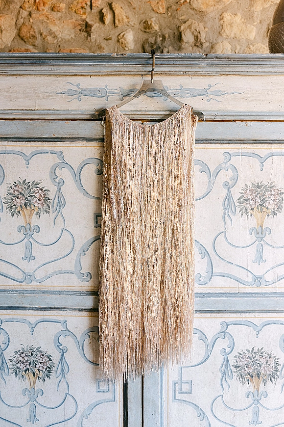 Wedding dress on hanger with beaded fringe detail, hanging against a painted vintage door with floral motifs and stone wall backdrop