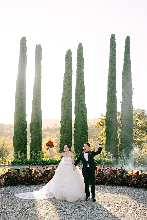 Newlywed portrait of bride holding bouquet up while holding hands with groom in tux, cheering on a cypress-lined garden terrace