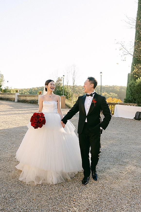 Couple portrait of bride and groom laughing, holding hands in a gravel courtyard; she wears a veil and carries a red rose bouquet