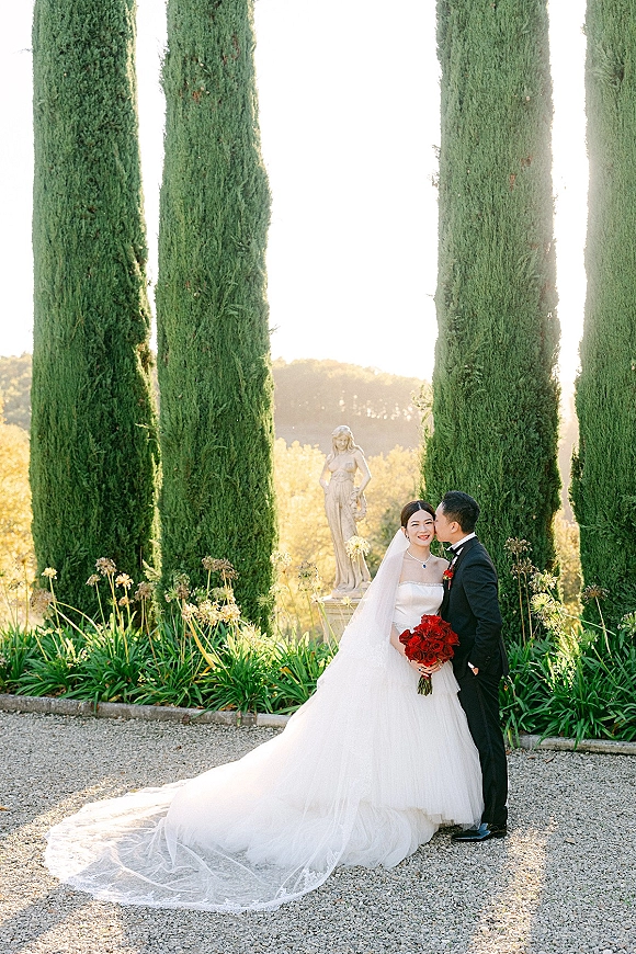 Couple portrait of groom kissing the bride’s cheek as she holds red roses, her long veil flowing along a garden path by cypress trees