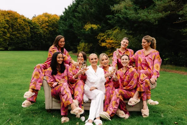 Bridesmaids getting ready in bridal party pajamas, sitting on a couch with the bride in a robe, raising champagne flutes on a lawn with trees