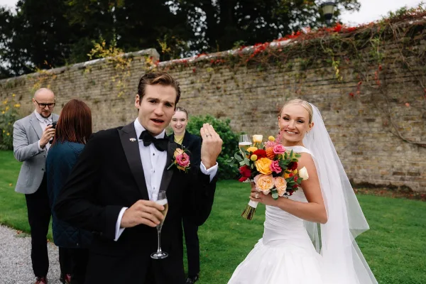 Wedding couple portrait of bride and groom candidly holding champagne as he shows his wedding ring, with bouquet accent by a stone wall