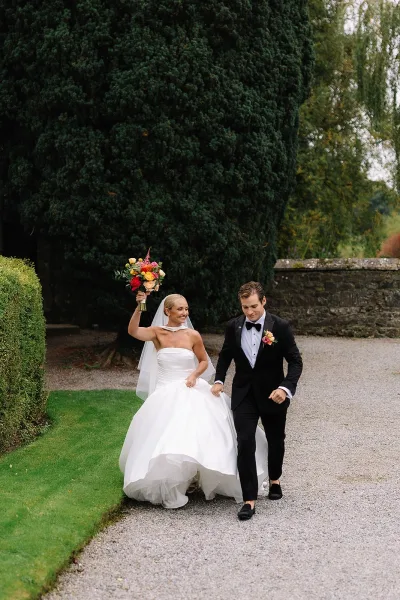 Recessional moment as bride and groom walking hand in hand, bride lifting bouquet, veil flowing on a gravel path in estate garden greenery