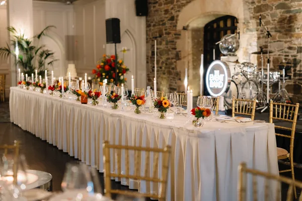 Reception head table with wedding sweetheart table decor, white linens, taper candles, bright florals, and gold chiavari chairs before a stage with neon band sign.