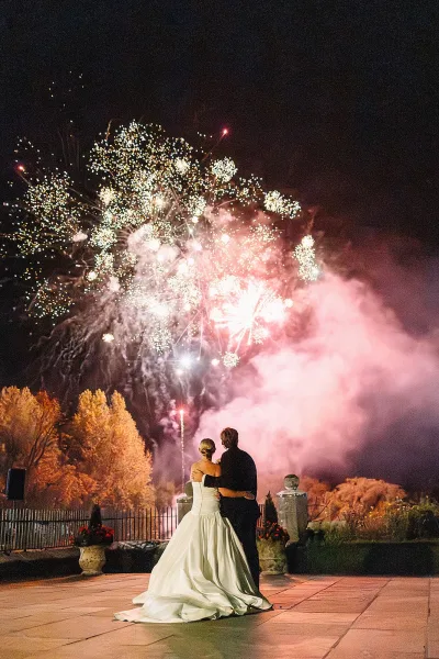 Wedding fireworks moment as bride and groom embrace from behind on a patio, watching bright fireworks burst through smoky night sky in the garden