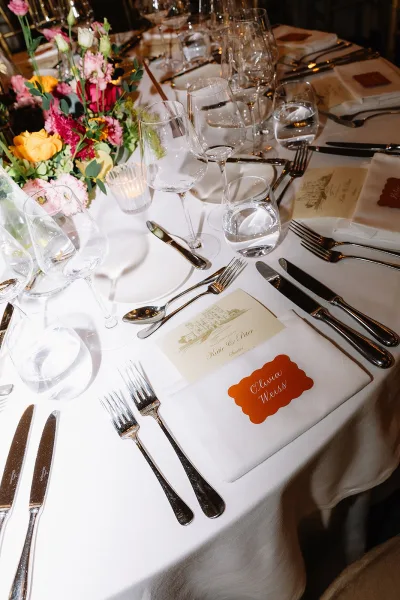 Reception tablescape with wedding place setting, white linen, floral rose centerpiece, orange place card, menu card, and votive candle in dim light