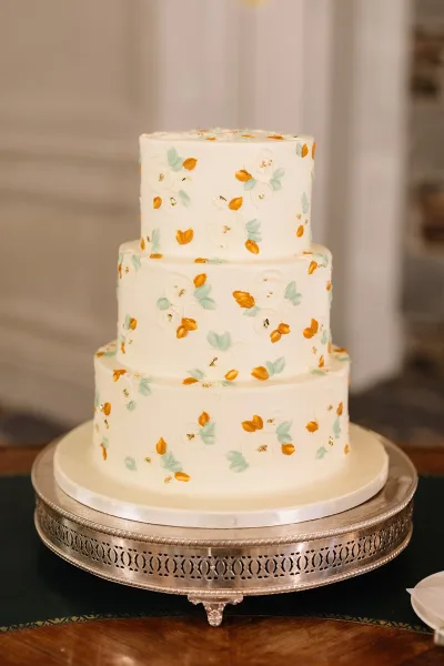 Wedding cake with white frosting and floral icing, three tier wedding cake with gold accents on a silver stand in a neutral room