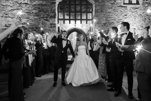 Sparkler exit as bride in strapless ball gown and groom in tuxedo walk through guest-lit tunnel by stone archway at night