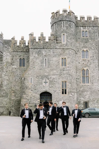 Groomsmen portrait of men in black tuxedos with bow ties holding beer glasses, walking in a stone castle courtyard under overcast sky