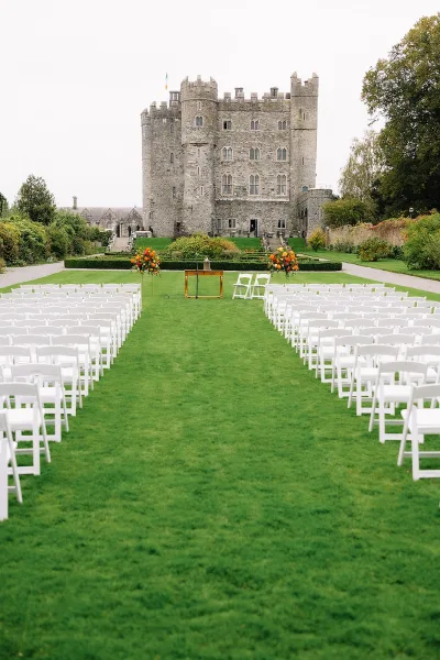 Outdoor ceremony setup with white folding chair rows framing a garden ceremony aisle, floral stands and mic before a stone castle lawn