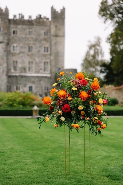 Ceremony floral arrangement of roses and dahlias with greenery on a metal stand, set on a lawn before a stone castle backdrop