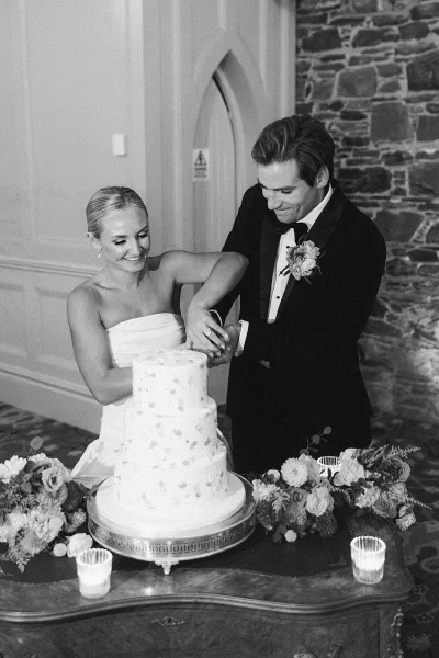 Cake cutting as bride in a strapless dress and groom in tuxedo slice a white two-tier wedding cake on a candlelit table by a stone arch