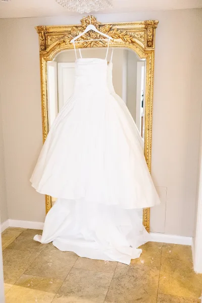 Wedding dress on a hanger reflected in an ornate gold mirror beneath a chandelier, set against a neutral wall by a doorway