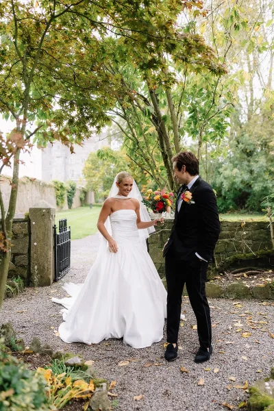 Couple portrait of bride in strapless dress and veil holding a colorful bouquet beside groom in black tuxedo on a garden gravel path