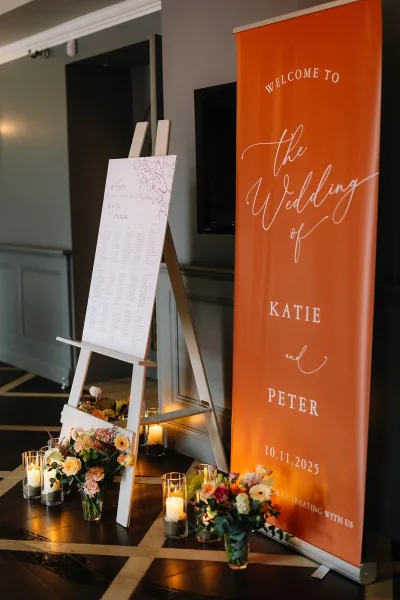 Wedding welcome sign with calligraphy lettering on an easel, flanked by florals and glass cylinder candles on a table by a paneled wall
