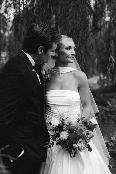 Couple portrait in a black and white wedding portrait, groom kissing bride’s cheek as she holds a bouquet under garden trees