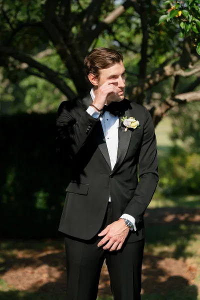 groom portrait capturing the groom wiping a tear, his white floral boutonniere crisp against his lapel, with garden trees and a neat hedge behind