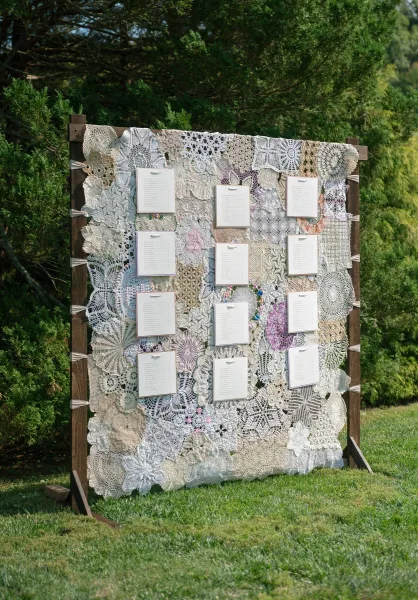 Wedding seating chart wall with guests’ names on lace doilies, set against garden trees and a sunlit grass lawn