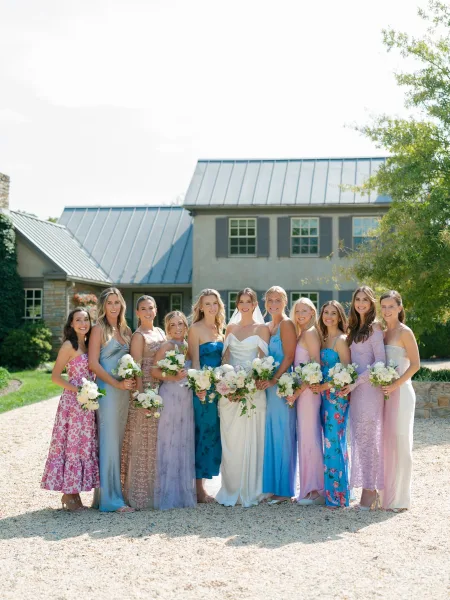 Bridal party portrait with mismatched bridesmaid dresses, the group holding bouquets on a gravel drive before a house framed by garden trees.