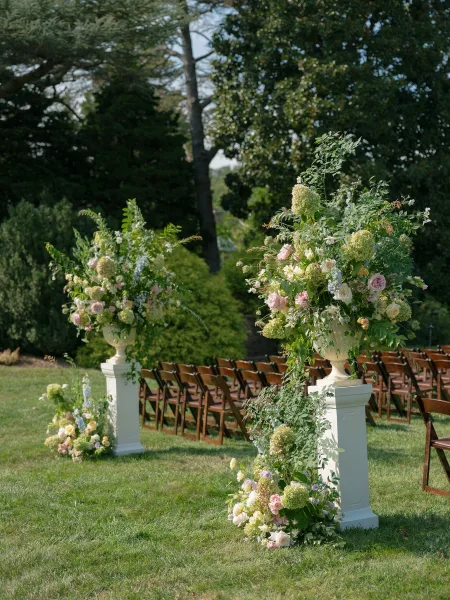 Ceremony aisle decor featuring outdoor ceremony flowers, with hydrangeas lining the aisle on a grass lawn framed by garden trees and open sky.