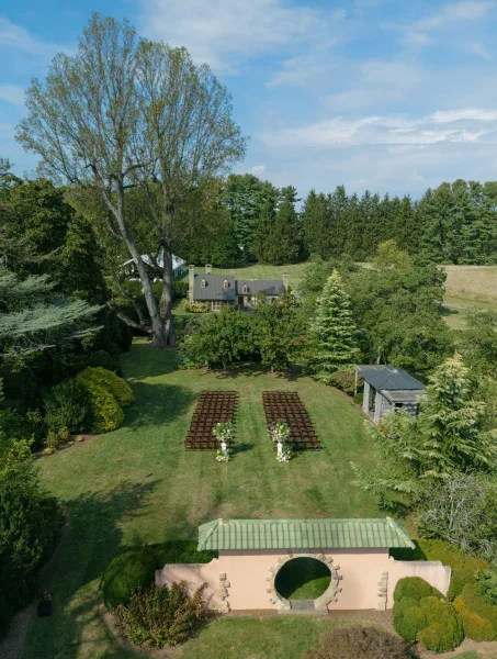 Outdoor ceremony setup with wooden chairs in a refined garden ceremony design, backed by garden trees and a cottage under a clear blue sky.