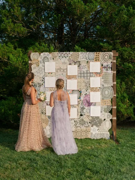 Seating chart display styled as a crochet lace seating chart, held in a wooden frame, set before garden trees and a grass lawn.