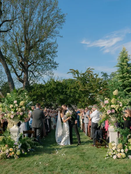 Recessional kiss during an outdoor ceremony exit, the bride clutching a bridal bouquet as garden trees and a blue sky frame their walk up the aisle.