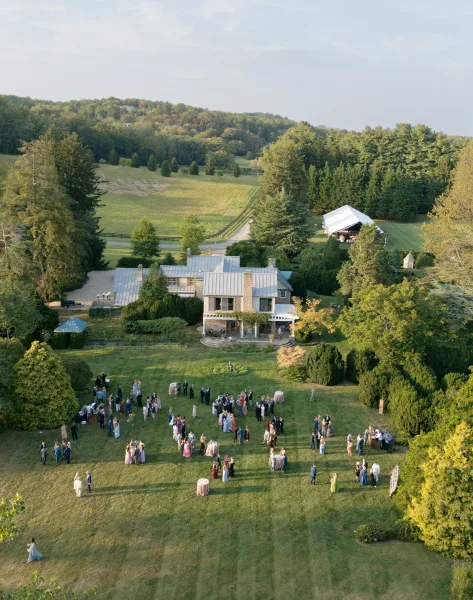 Wedding cocktail hour during an outdoor drinks reception on a grass lawn, with scattered cocktail tables, estate house and rolling hills beyond.