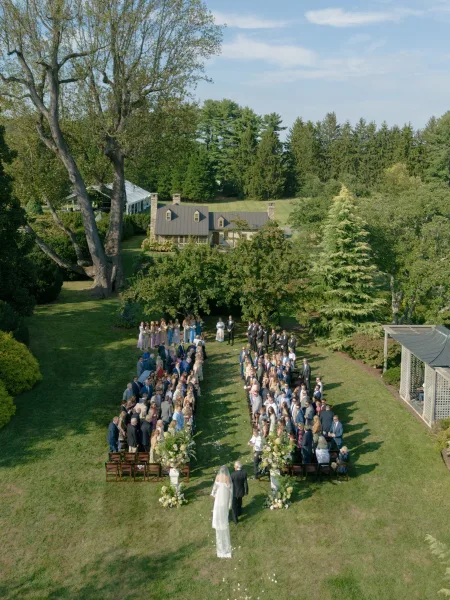 Wedding processional with a bride walking down the aisle in a long bridal veil on a sunlit garden lawn beneath blue sky and trees