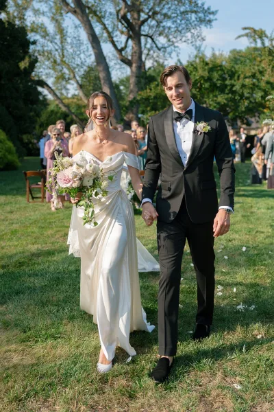 Wedding recessional with bride and groom walking, her blush and white bouquet with trailing vines as they cross a garden lawn past chairs.