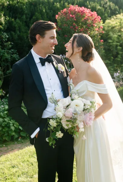 Couple portrait of a bride and groom laughing amid flowering garden greenery, as she holds a blush and white bridal bouquet.