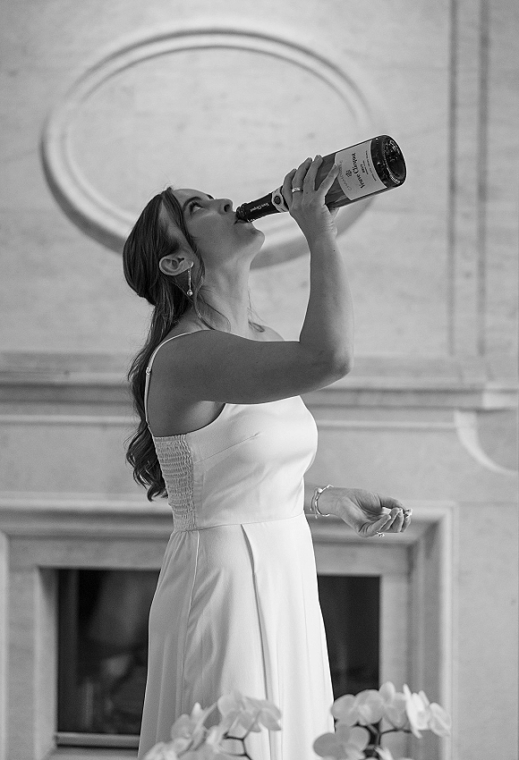 Bride drinking champagne beside a fireplace, holding a champagne bottle in her wedding dress with drop earrings, bracelet, and rings