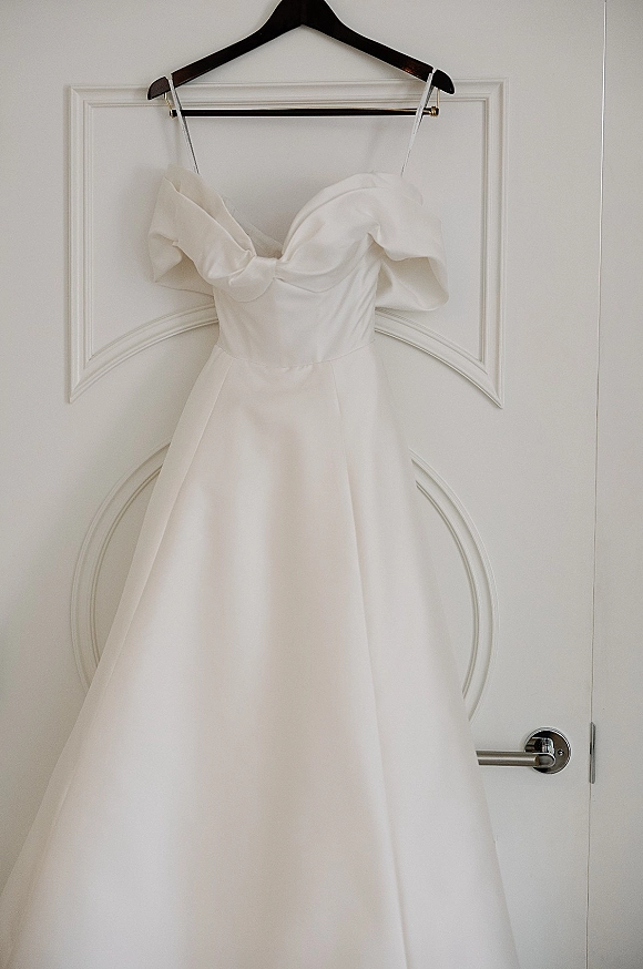Wedding dress with off the shoulder sleeves hanging on a black hanger against a white door with molding and handle