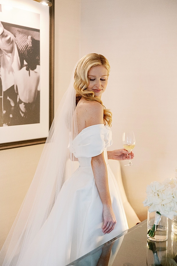 Bridal portrait of a blonde bride in an off the shoulder wedding dress with long veil, holding a wine glass beside a table with framed art in warm light