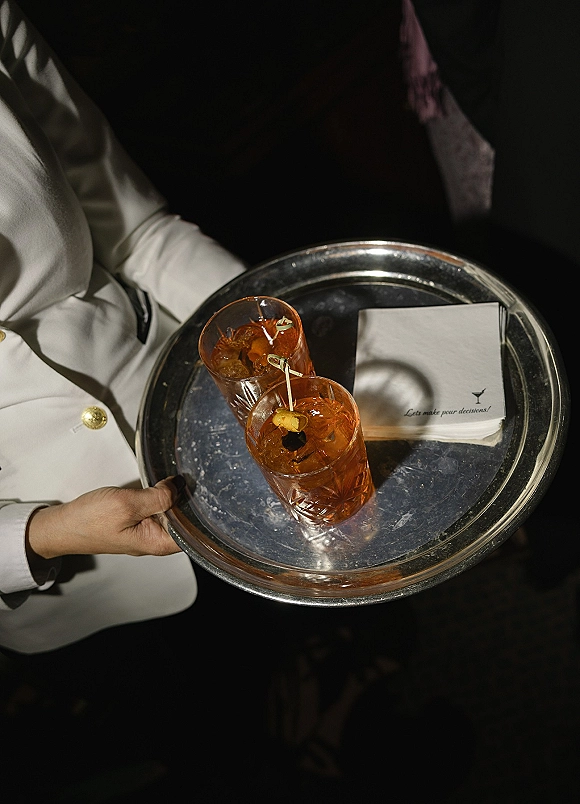 Wedding cocktails on a silver serving tray with ice-filled glasses, olive and orange garnishes, held by a server in a dark indoor setting