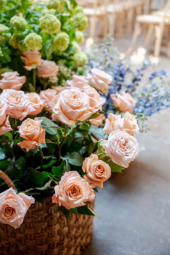 Wedding floral arrangement with peach rose wedding flowers in a woven basket, featuring hydrangeas and blue blooms beside wooden chairs indoors
