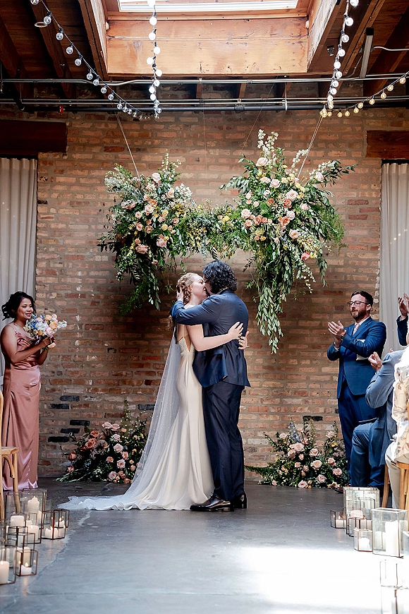 Wedding kiss under a floral ceremony arch with greenery and roses, string lights and candles glowing against an exposed brick loft backdrop