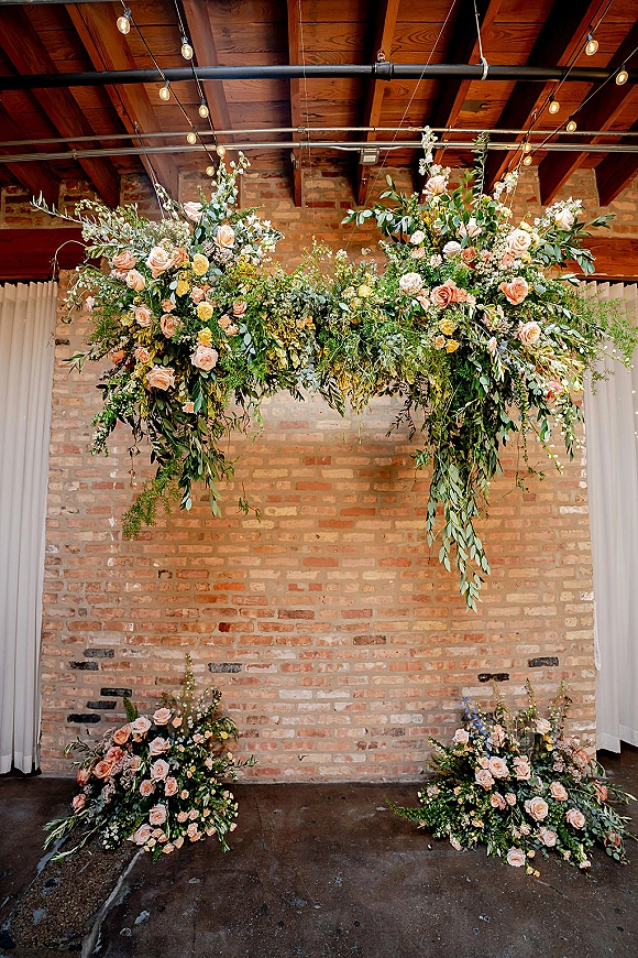 Wedding floral installation with hanging greenery and blush, peach, and white roses beneath string lights against an industrial brick wall backdrop