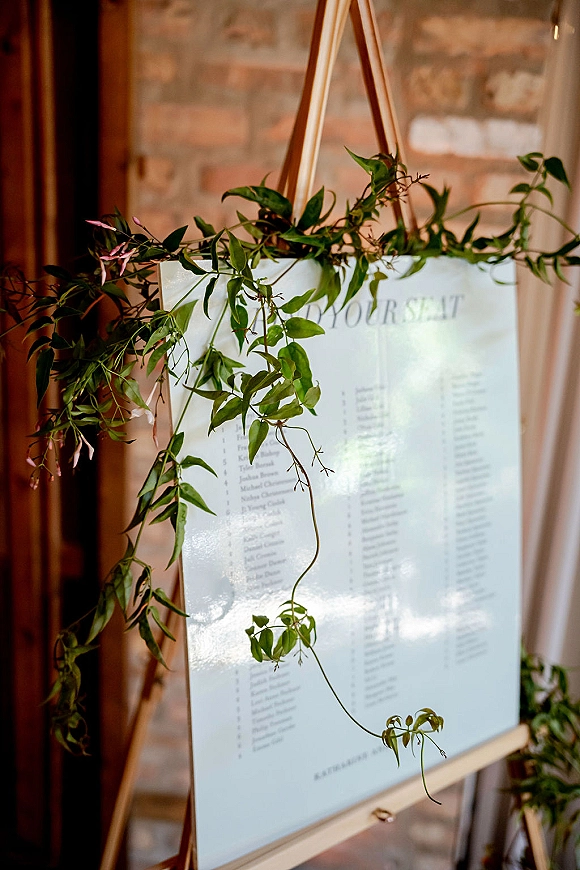 Wedding seating chart displayed on a wood easel with greenery garland, set against a brick wall inside the venue