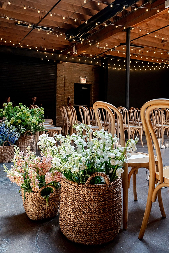 Ceremony setup with indoor ceremony seating on bistro chairs, candlelit aisle runner and floral baskets under string lights in an industrial loft