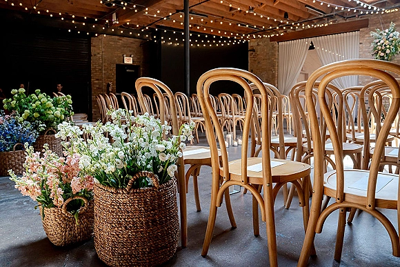 Ceremony seating with bentwood chair rows under string lights, floral baskets and white draping against a brick wall in an industrial venue