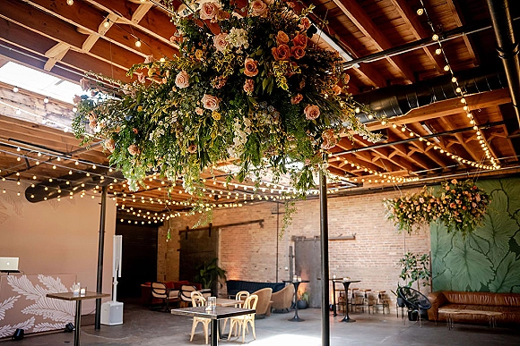 Hanging floral installation with blush roses and greenery under string lights, floating above an industrial loft lounge with brick walls