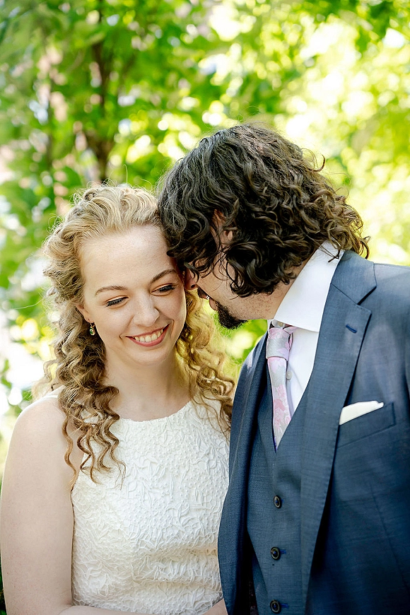 Couple portrait of bride and groom touching foreheads as he whispers, her lace dress in sunlight with green trees behind