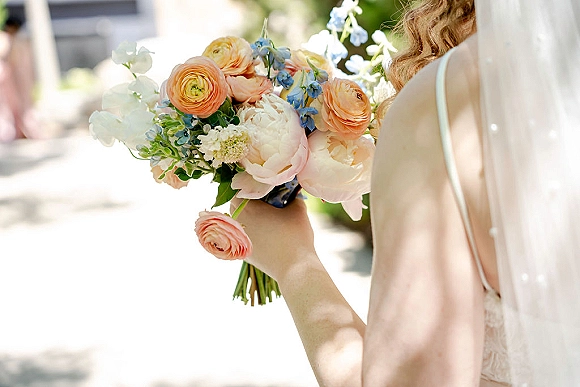 Bridal bouquet of peach ranunculus and blush peonies with blue delphinium and greenery, held by bride in veil on sunlit walkway