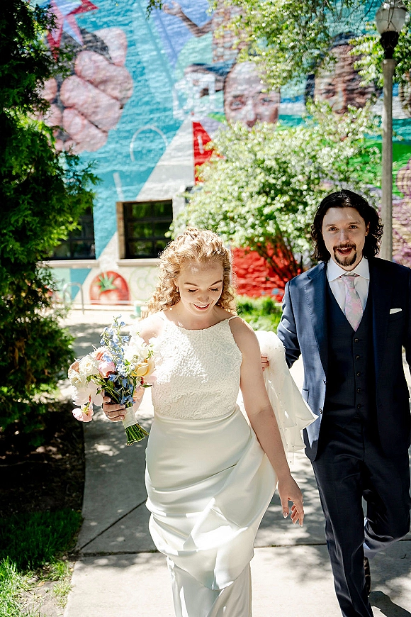 Couple portrait of bride and groom walking, bride holding bouquet with blue flowers, passing a colorful mural on a city sidewalk